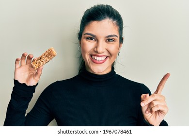 Young Hispanic Woman Eating Protein Bar As Healthy Energy Snack Smiling Happy Pointing With Hand And Finger To The Side 