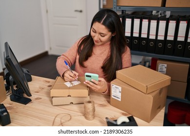 Young Hispanic Woman Ecommerce Business Worker Writing On Package And Using Smartphone At Office