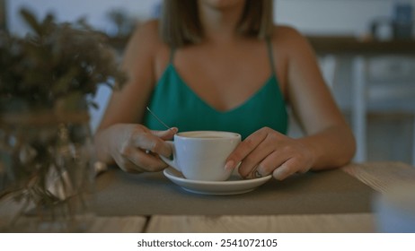 Young hispanic woman enjoys coffee indoors at a cozy cafeteria in italy, wearing green top and nail polish, holding cup with both hands. - Powered by Shutterstock