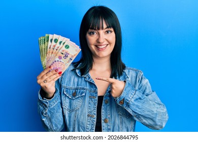 Young hispanic woman holding argentine pesos banknotes smiling happy pointing with hand and finger 
