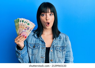 Young hispanic woman holding argentine pesos banknotes scared and amazed with open mouth for surprise, disbelief face 