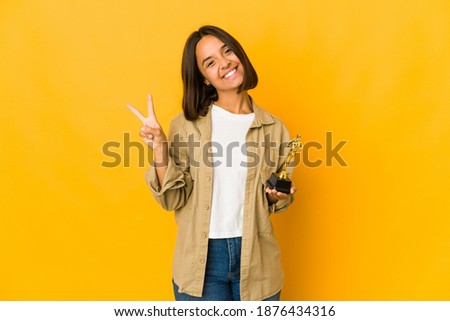 Young hispanic woman holding an award statuette joyful and carefree showing a peace symbol with fingers.