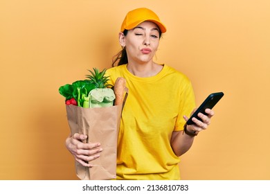 Young Hispanic Woman Holding Bag Of Groceries Using Smartphone Looking At The Camera Blowing A Kiss Being Lovely And Sexy. Love Expression. 