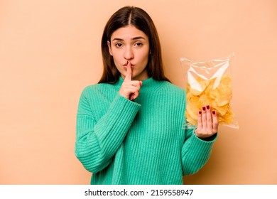 Young Hispanic Woman Holding A Bag Of Chips Isolated On Beige Background Keeping A Secret Or Asking For Silence.