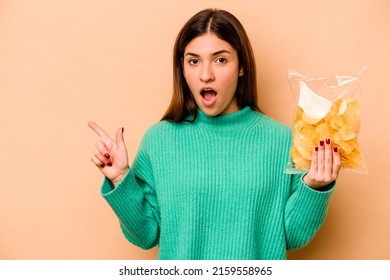 Young Hispanic Woman Holding A Bag Of Chips Isolated On Beige Background Pointing To The Side