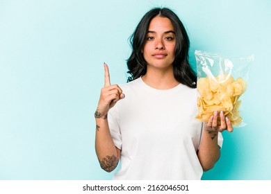 Young Hispanic Woman Holding A Bag Of Chips Isolated On Blue Background Showing Number One With Finger.