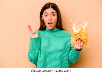 Young Hispanic Woman Holding A Bag Of Chips Isolated On Beige Background Surprised And Shocked.