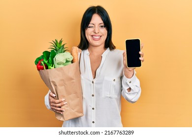 Young Hispanic Woman Holding Bag Of Groceries Using Smartphone Winking Looking At The Camera With Sexy Expression, Cheerful And Happy Face. 