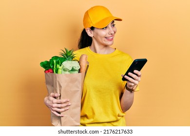 Young Hispanic Woman Holding Bag Of Groceries Using Smartphone Smiling Looking To The Side And Staring Away Thinking. 