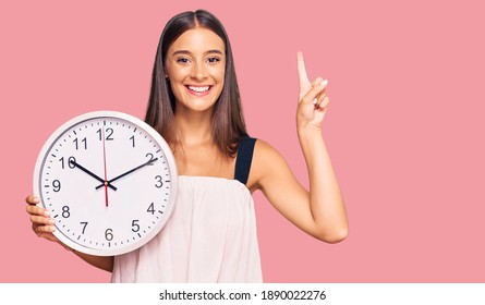 Young Hispanic Woman Holding Big Clock Surprised With An Idea Or Question Pointing Finger With Happy Face, Number One 