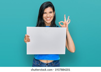 Young hispanic woman holding blank empty banner doing ok sign with fingers, smiling friendly gesturing excellent symbol 