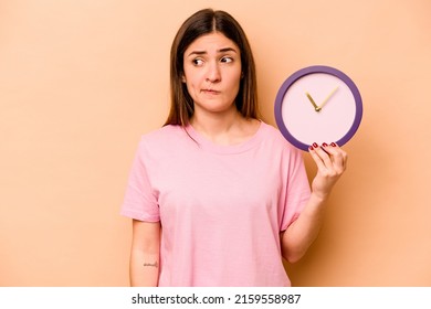Young hispanic woman holding a clock isolated on beige background confused, feels doubtful and unsure.