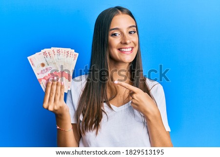Young hispanic woman holding colombian pesos smiling happy pointing with hand and finger 