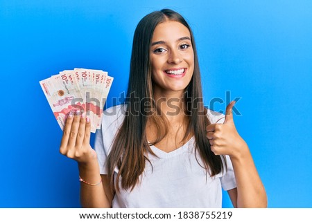Young hispanic woman holding colombian pesos smiling happy and positive, thumb up doing excellent and approval sign 