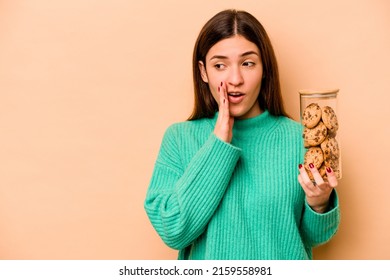 Young Hispanic Woman Holding Cookies Jar Isolated On Beige Background Is Saying A Secret Hot Braking News And Looking Aside