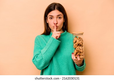 Young Hispanic Woman Holding Cookies Jar Isolated On Beige Background Keeping A Secret Or Asking For Silence.