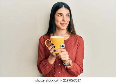 Young hispanic woman holding cup of tea smiling looking to the side and staring away thinking. 