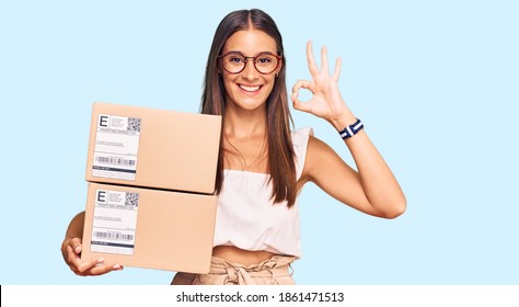 Young hispanic woman holding delivery package doing ok sign with fingers, smiling friendly gesturing excellent symbol 