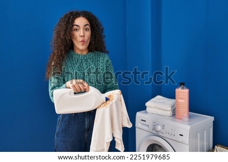 Young hispanic woman holding dirty laundry and detergent bottle making fish face with mouth and squinting eyes, crazy and comical. 
