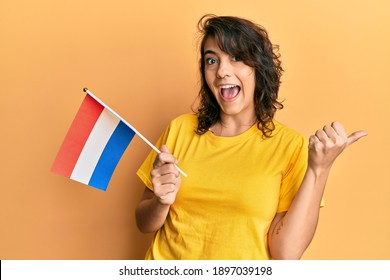 Young hispanic woman holding holland flag pointing thumb up to the side smiling happy with open mouth 