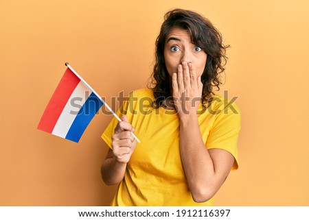 Young hispanic woman holding holland flag covering mouth with hand, shocked and afraid for mistake. surprised expression 