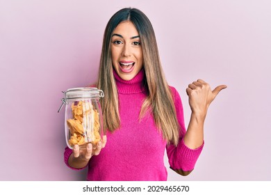 Young hispanic woman holding jar of uncooked pasta pointing thumb up to the side smiling happy with open mouth 