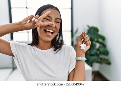 Young Hispanic Woman Holding Keys Of New Home Doing Peace Symbol With Fingers Over Face, Smiling Cheerful Showing Victory 