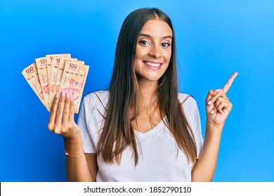 Young hispanic woman holding mexican pesos smiling happy pointing with hand and finger to the side 