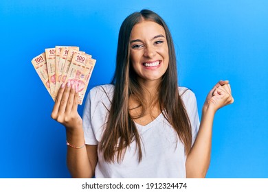 Young Hispanic Woman Holding Mexican Pesos Screaming Proud, Celebrating Victory And Success Very Excited With Raised Arm 