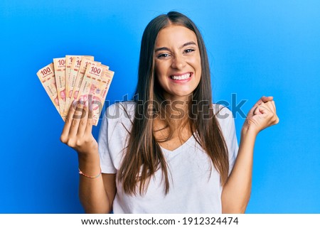 Young hispanic woman holding mexican pesos screaming proud, celebrating victory and success very excited with raised arm 