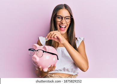 Young hispanic woman holding piggy bank with glasses and coin smiling and laughing hard out loud because funny crazy joke. 