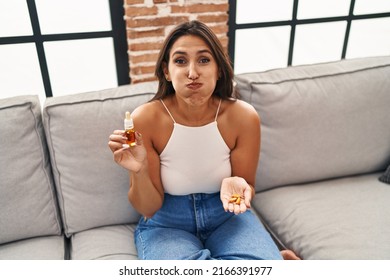 Young Hispanic Woman Holding Pills And Cbd Oil Puffing Cheeks With Funny Face. Mouth Inflated With Air, Catching Air. 