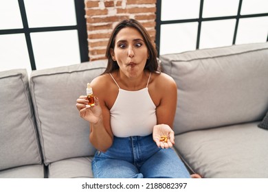 Young Hispanic Woman Holding Pills And Cbd Oil Making Fish Face With Mouth And Squinting Eyes, Crazy And Comical. 