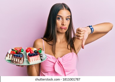 Young Hispanic Woman Holding Plate With Cheesecake With Angry Face, Negative Sign Showing Dislike With Thumbs Down, Rejection Concept 