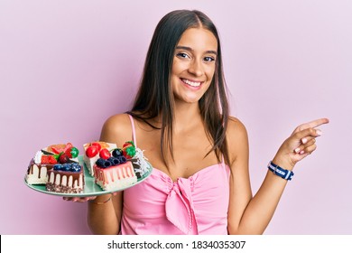 Young Hispanic Woman Holding Plate With Cheesecake Smiling Happy Pointing With Hand And Finger To The Side 