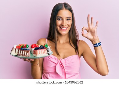 Young Hispanic Woman Holding Plate With Cheesecake Doing Ok Sign With Fingers, Smiling Friendly Gesturing Excellent Symbol 