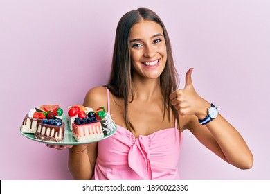 Young Hispanic Woman Holding Plate With Cheesecake Smiling Happy And Positive, Thumb Up Doing Excellent And Approval Sign 