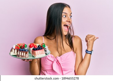 Young Hispanic Woman Holding Plate With Cheesecake Pointing Thumb Up To The Side Smiling Happy With Open Mouth 