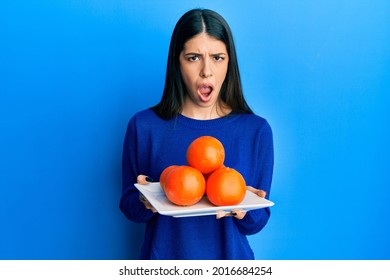 Young Hispanic Woman Holding Plate With Fresh Oranges Clueless And Confused Expression. Doubt Concept. 