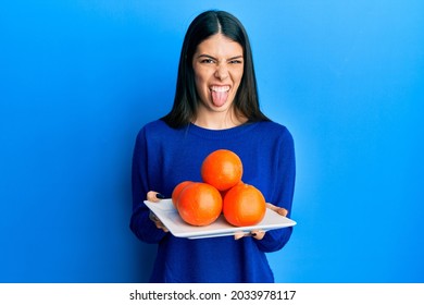 Young Hispanic Woman Holding Plate With Fresh Oranges Sticking Tongue Out Happy With Funny Expression. 