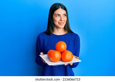 Young Hispanic Woman Holding Plate With Fresh Oranges Smiling Looking To The Side And Staring Away Thinking. 