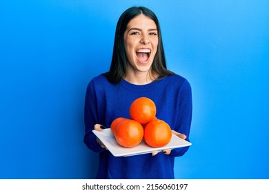 Young Hispanic Woman Holding Plate With Fresh Oranges Smiling And Laughing Hard Out Loud Because Funny Crazy Joke. 