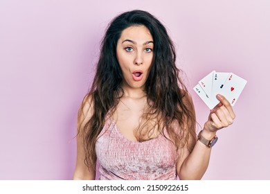 Young Hispanic Woman Holding Poker Cards Scared And Amazed With Open Mouth For Surprise, Disbelief Face 