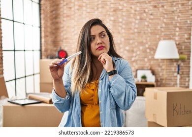 Young Hispanic Woman Holding Pregnancy Test Result At New Home Serious Face Thinking About Question With Hand On Chin, Thoughtful About Confusing Idea 