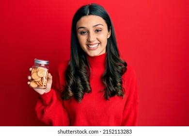 Young hispanic woman holding salty biscuits jar looking positive and happy standing and smiling with a confident smile showing teeth 