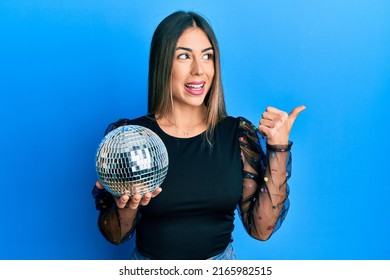 Young hispanic woman holding shiny disco ball pointing thumb up to the side smiling happy with open mouth 