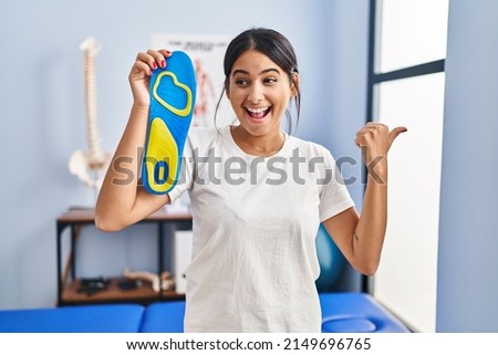 Young hispanic woman holding shoe insole at physiotherapy clinic pointing thumb up to the side smiling happy with open mouth 