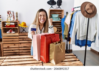 Young Hispanic Woman Holding Shopping Bags And Credit Card At Clothing Store Angry And Mad Raising Fist Frustrated And Furious While Shouting With Anger. Rage And Aggressive Concept. 