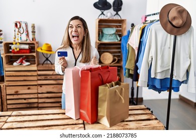 Young Hispanic Woman Holding Shopping Bags And Credit Card At Clothing Store Angry And Mad Screaming Frustrated And Furious, Shouting With Anger. Rage And Aggressive Concept. 