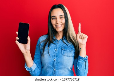 Young Hispanic Woman Holding Smartphone Showing Screen Smiling With An Idea Or Question Pointing Finger With Happy Face, Number One 